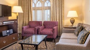Ein Hotelzimmer mit Flachbildfernseher, einer Holzkassette mit DVD-Player, zwei gemusterten Sesseln, einem Glastisch und einer Couch mit gemusterten Kissen sowie einem Fenster mit Vorhängen.