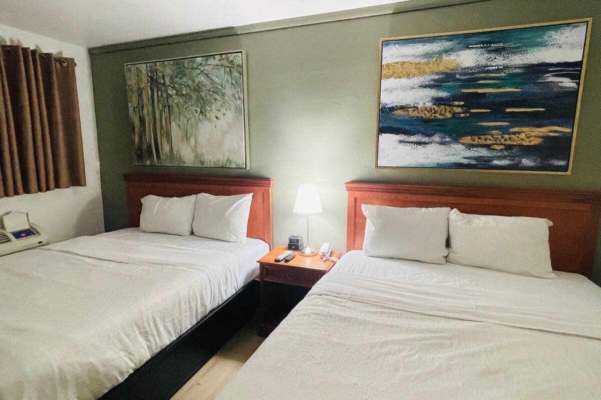 Habitación de hotel con dos camas, una mesita de noche con lámpara, un cuadro de un bosque y un cuadro con una playa.