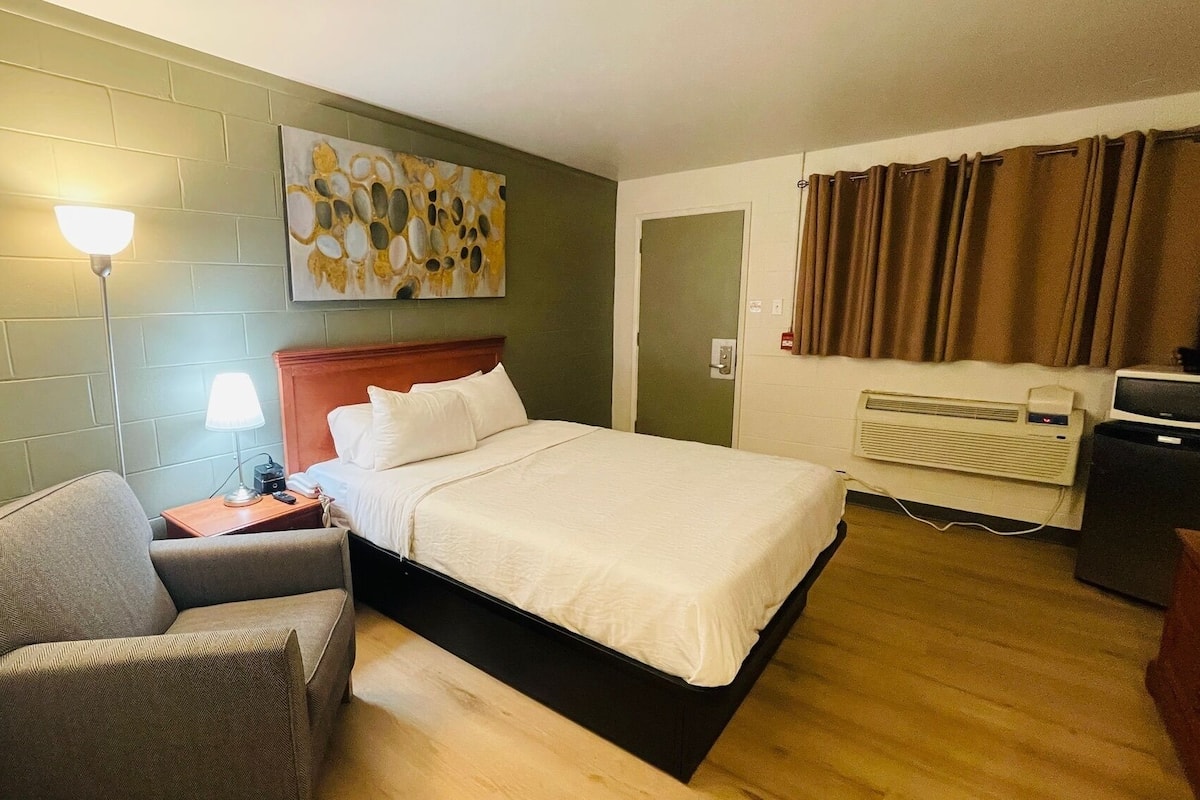 Habitación de hotel con cama, una silla, mesita de noche, lámpara, un cuadro, calentador y ventana con cortinas.