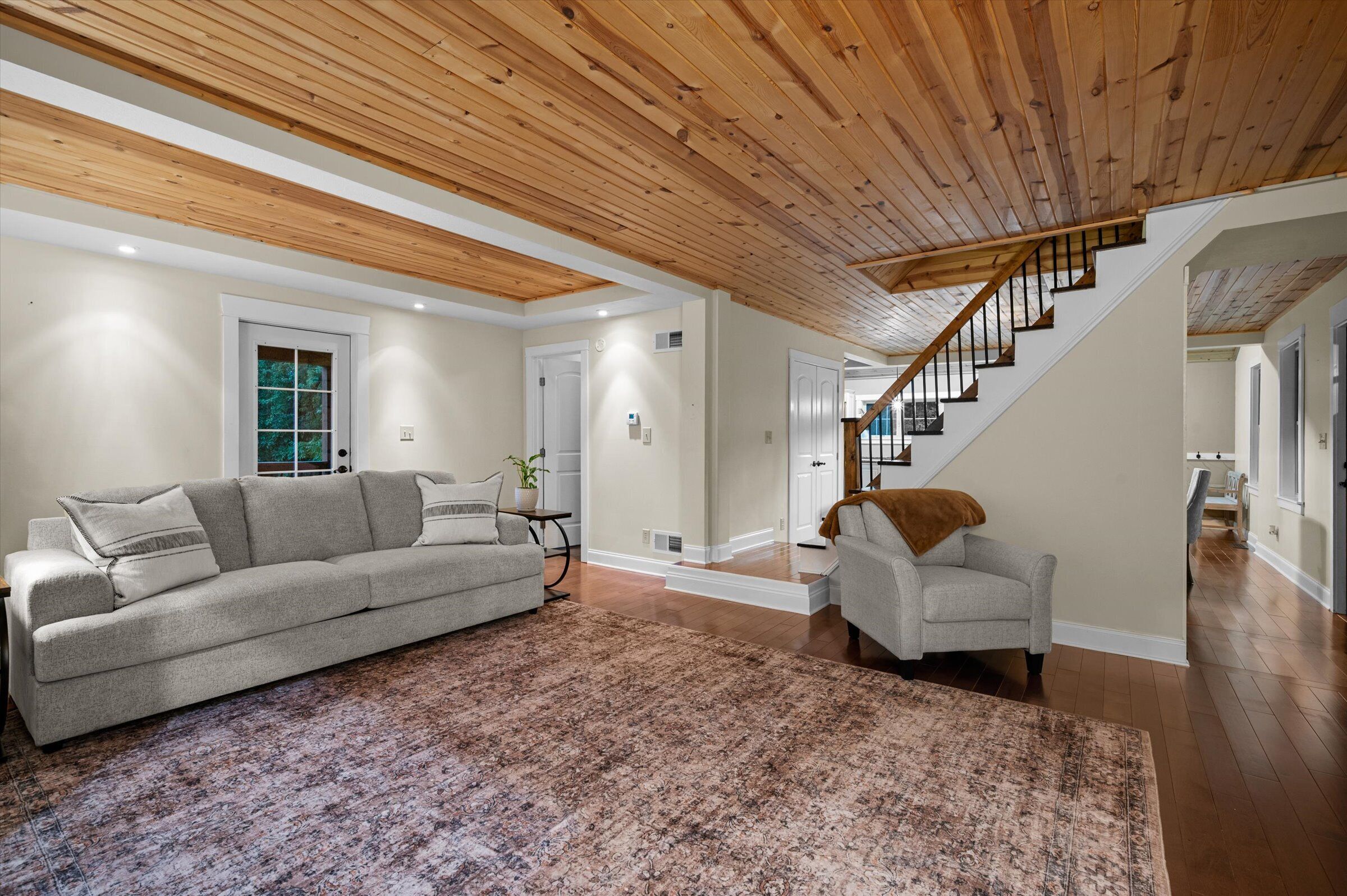 A spacious living room with a large gray sofa, a smaller armchair, and a patterned rug. A wooden staircase with a railing leads to another level.