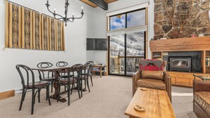 A cozy living room with a fireplace, a dining area with chairs and a table, and a large window offering a view of a snowy landscape. - Mountain Views | Close to Slopes | Hot Tubs (steamboat springs)