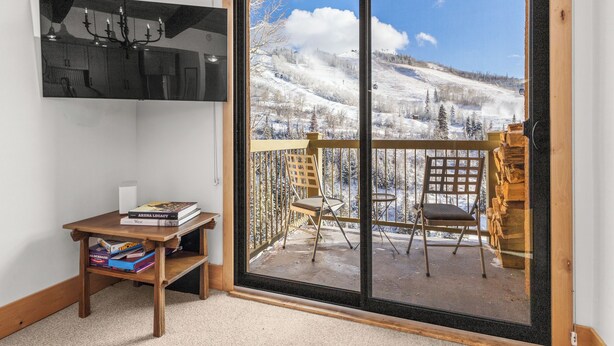 A room with a wooden table, books, and a large window with a view of a snowy mountain. - Mountain Views | Close to Slopes | Hot Tubs (steamboat springs)