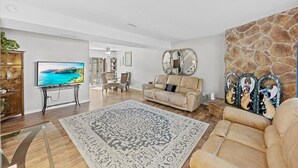 Ein geräumiges Wohnzimmer mit einem großen Fernseher, einem beigefarbenen Sofa, einem gemusterten Teppich und einer Wand mit Steinverkleidung.