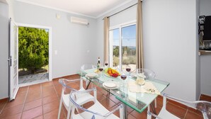 Zona pranzo con tavolo in vetro, sedie bianche e vista su vegetazione e un paesaggio lontano attraverso ampie finestre.