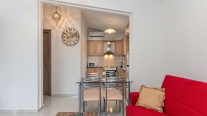 Un salon moderne avec un canapé rouge, une table à manger en verre entourée de chaises, une table basse en bois et, au fond, une cuisine dotée de meubles en bois et d’un four à micro-ondes.
