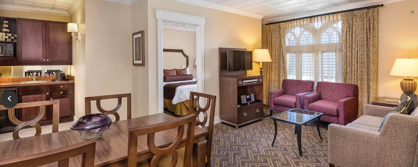 A hotel room with a dining area, a living room with a sofa and armchairs, a TV, and a window with curtains.