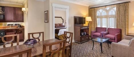 A hotel room with a dining area, a living room with a sofa and armchairs, a TV, and a window with curtains.