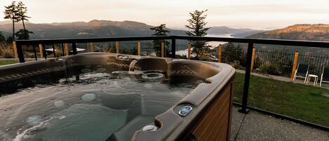 Een hottub met een houten buitenkant, omringd door een metalen reling, met uitzicht op een schilderachtig berglandschap en een waterpartij.