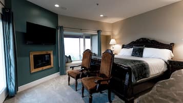 Un dormitorio con una cama grande, dos sillas, un televisor montado en la pared y un ventanal con cortinas.