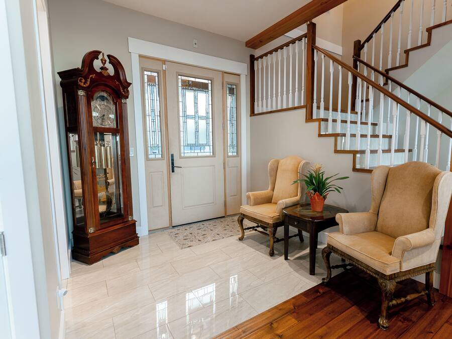 A spacious living area with a wooden staircase, two armchairs, a large grandfather clock, and a potted plant.