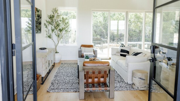 A modern living room with a white sofa, a wooden bench, and a glass coffee table. There is a large window providing a view of the outdoors. - Luxury Ranch Near Palo Pinto Park with 10 Beds (Strawn)