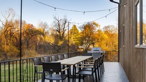 Un patio al aire libre con una larga mesa de madera y sillas, una parrilla y vistas a árboles otoñales.
