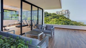 Een modern balkon met buitenmeubilair, een houten vloer, grijs en wit meubilair en een uitzicht op een weelderige groene heuvel en gebouwen in de verte.