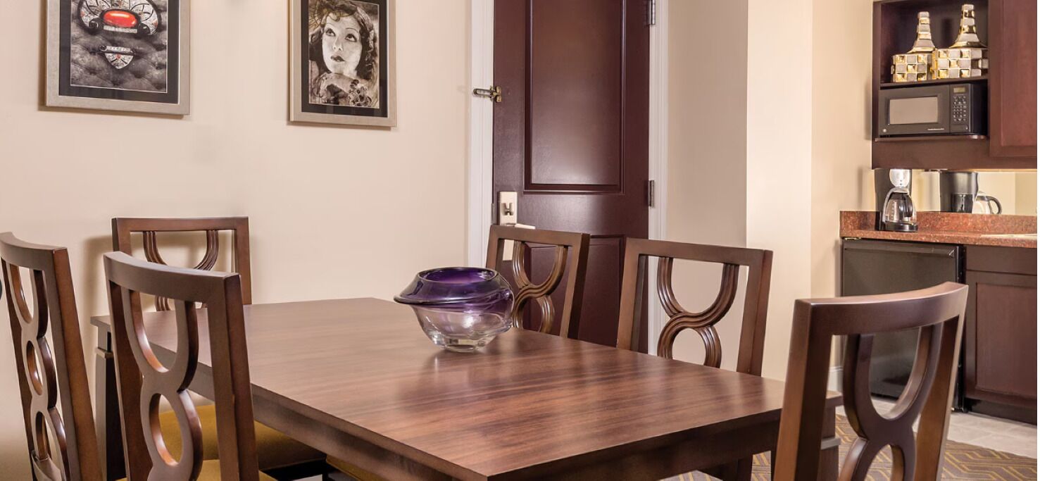 A dining area with a wooden table and chairs, a purple bowl, framed artwork on the wall, and a kitchen area with appliances.