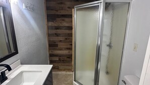 Une salle de bain avec un lavabo blanc, des toilettes, une douche en verre et un mur en bois.