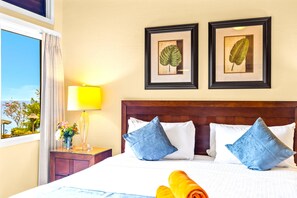 Ein Hotelzimmer mit einem großen Bett, zwei gerahmten Pflanzenbildern an der Wand, einer Nachttischlampe und Blick ins Freie.