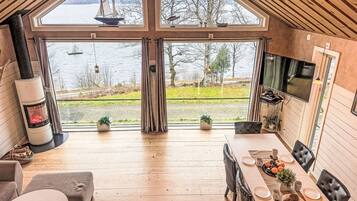 Uma cabana acolhedora com uma grande janela com vista para um lago, lareira e área de jantar com TV.