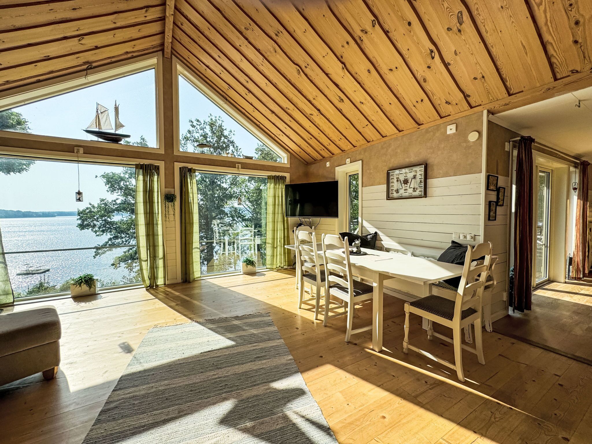 A spacious room with a wooden ceiling, large windows offering a view of a lake, a dining area with a table and chairs, and a cozy seating area with a sofa.