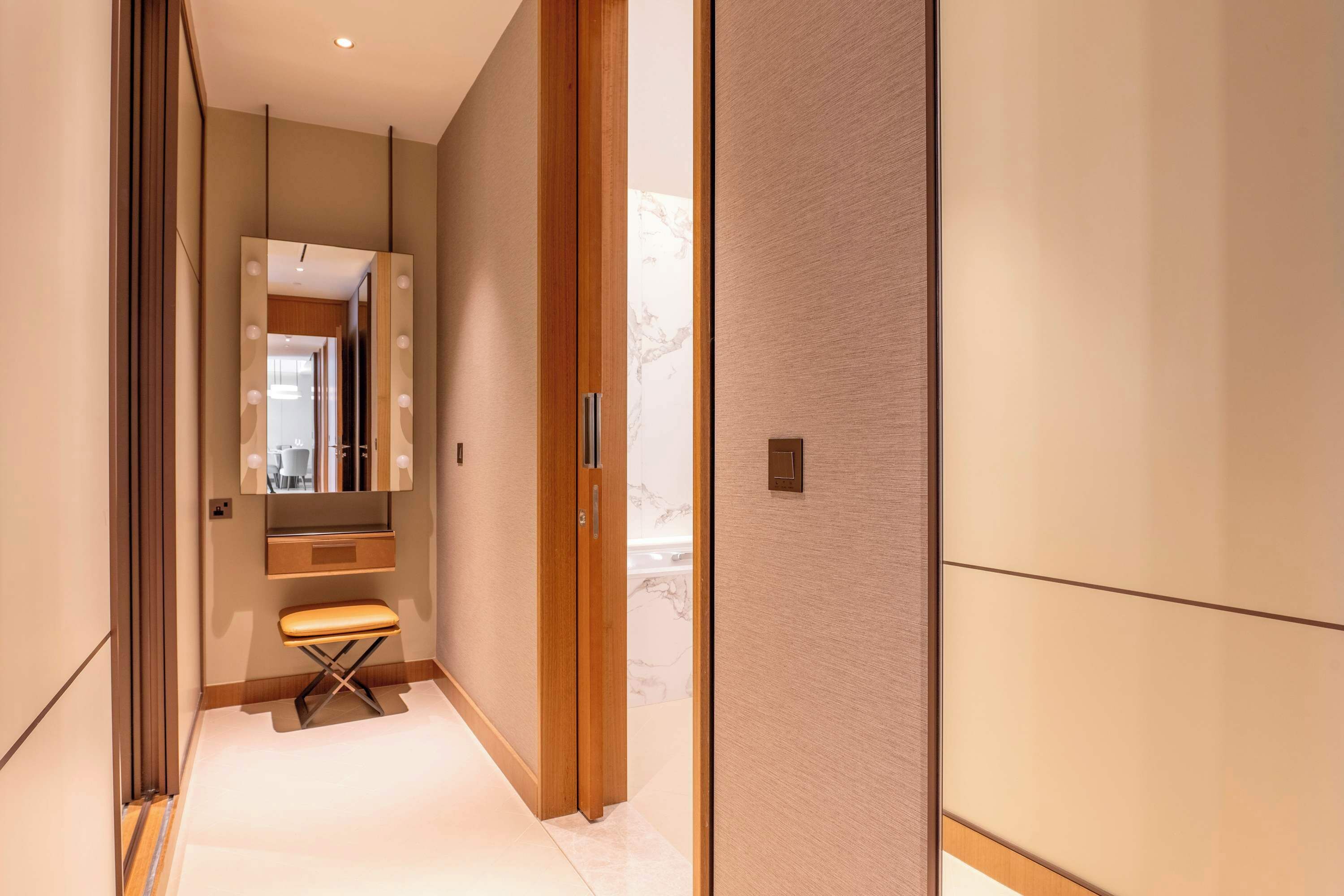 Un couloir moderne avec un banc, un miroir muni de lampes et une porte donnant sur une pièce.
