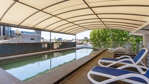 Un área de piscina en la azotea con toldo, sillas de descanso y vistas de la ciudad.