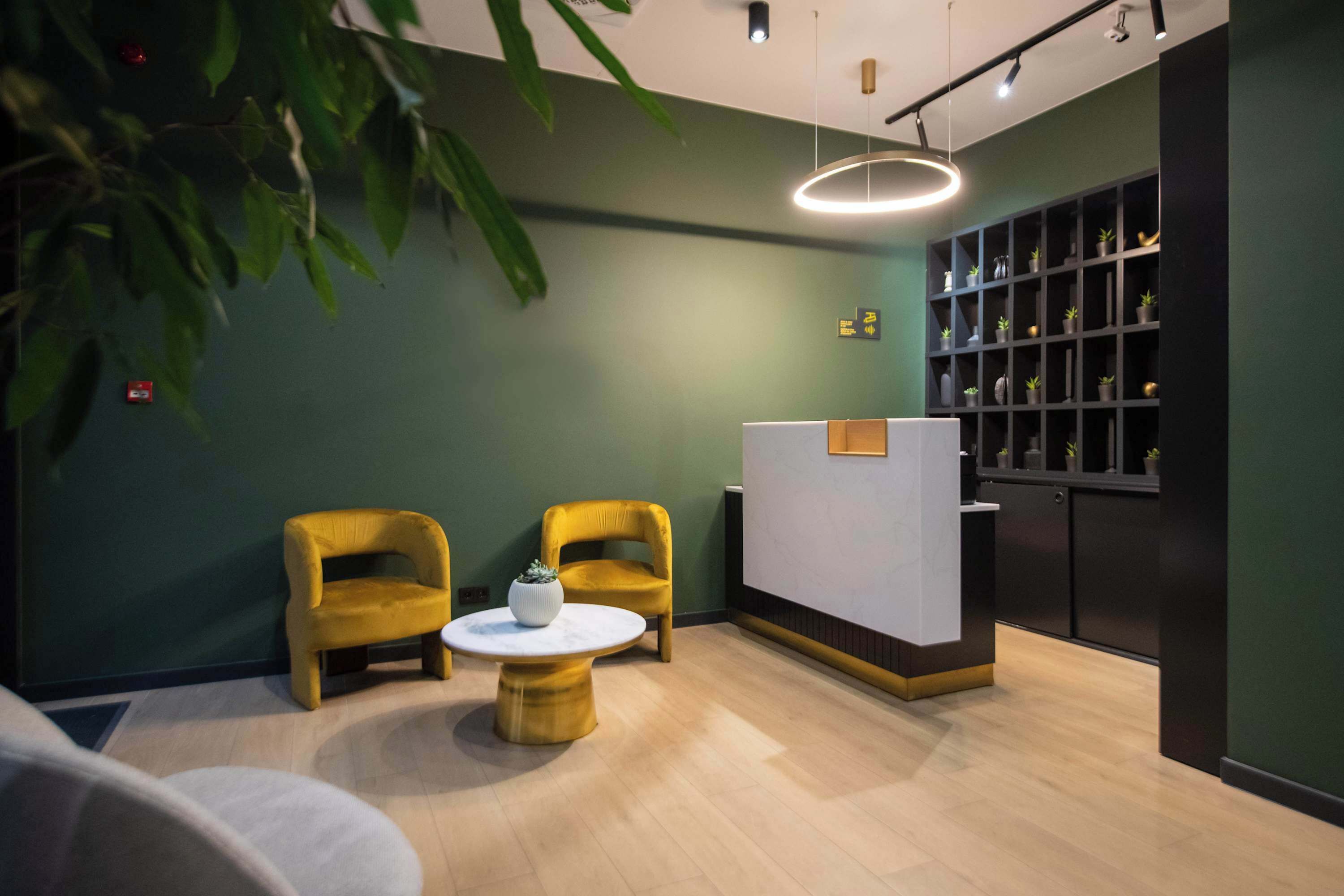 A reception area with a reception desk, two yellow chairs, a round table with a white vase, and a plant.
