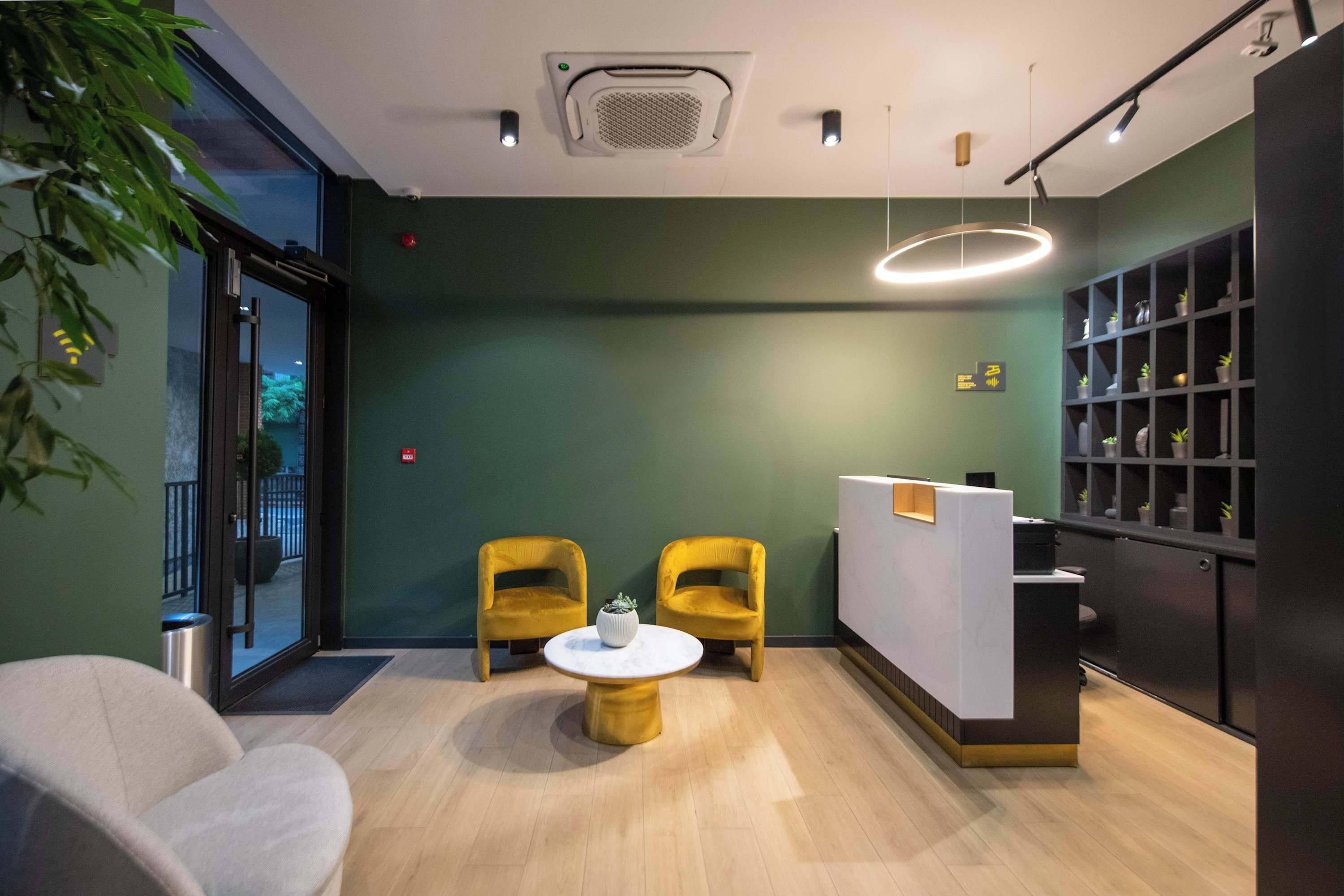A reception area with a reception desk, two yellow chairs, a round table with a white vase, and a green wall with a shelf of plants.