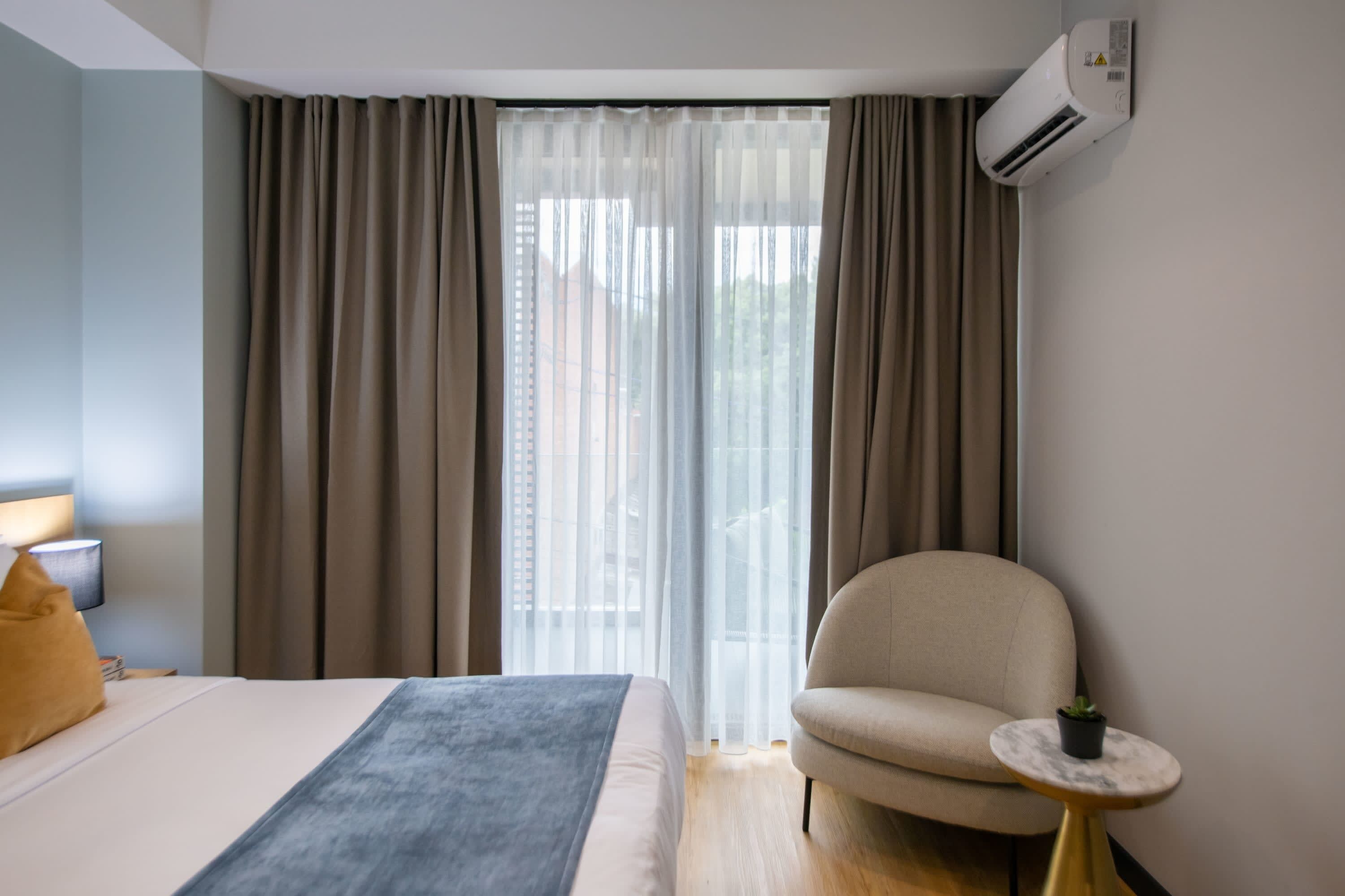 A hotel room with a bed, a chair, a small table with a plant, and an air conditioning unit.