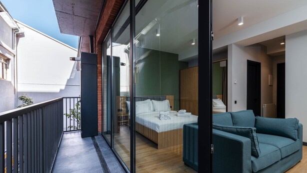 A modern hotel room with a balcony, a bed, a sofa, and a view of the city. - Studio Balcony Suite with Kitchen (T'bilisi)