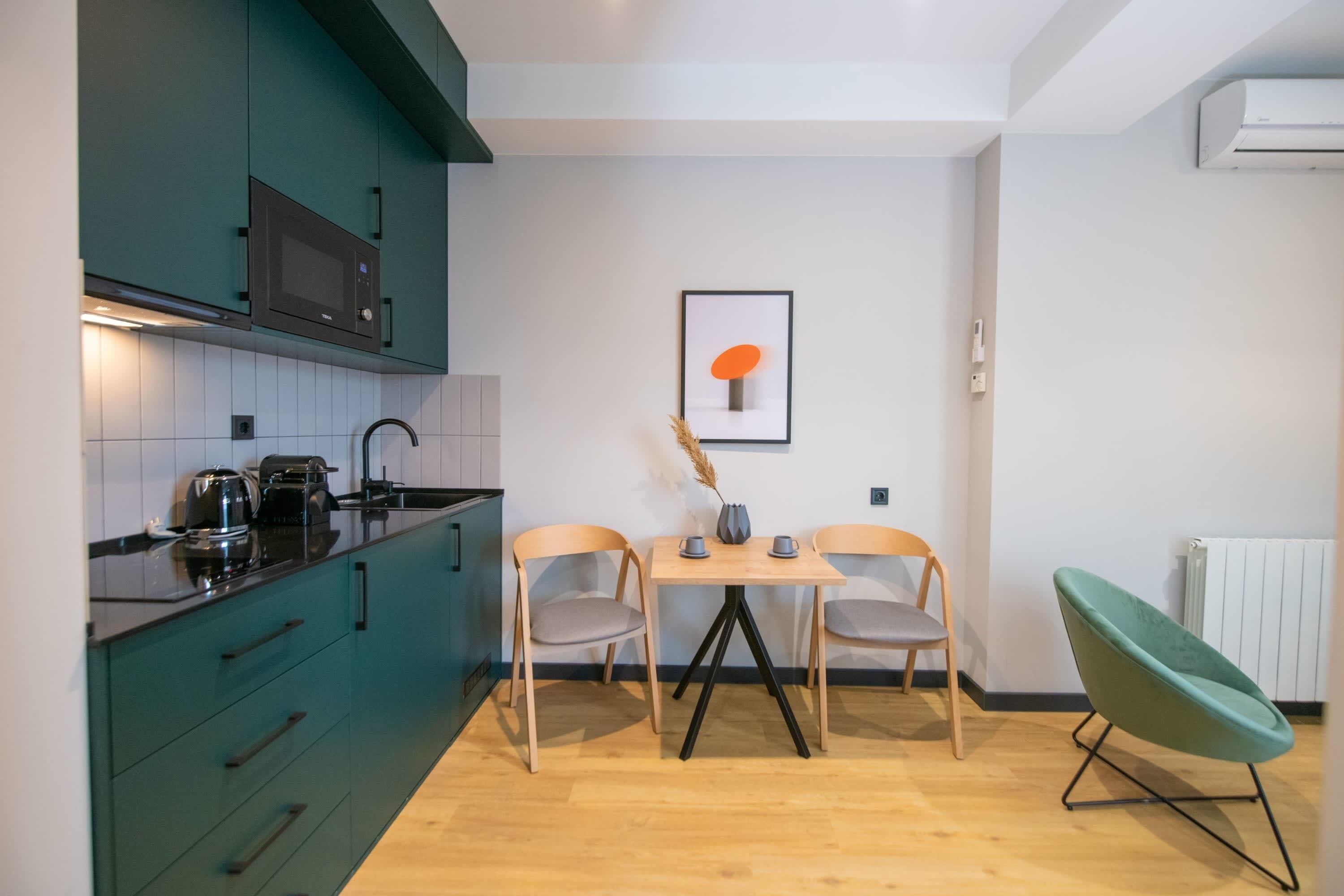 Cocina compacta con armarios verde azulado, un microondas y una pequeña mesa de comedor con dos sillas.