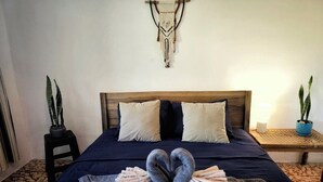 A bed with a dark blue bedspread, two pillows, and a decorative macrame wall hanging. There are two potted plants, one on a small table and another on a stool. - 4 Amigos - Seaview Room OLITA, 5 min to the beach, pool, private bathroom (El Zonte)