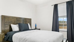 A bedroom with a wooden headboard, a bed with white bedding, a nightstand, a blue lamp, and a window with black curtains.