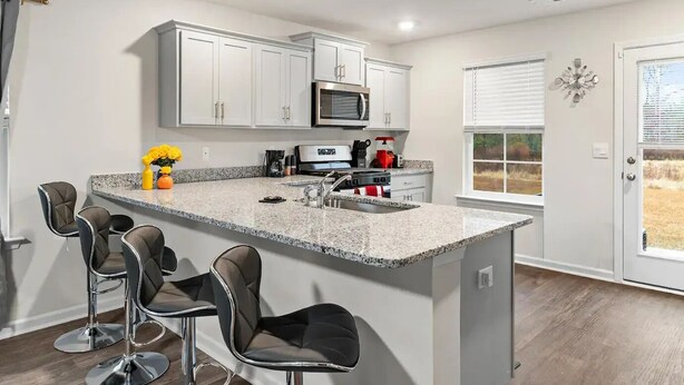 A modern kitchen with white cabinets, a granite countertop, and stainless steel appliances. There are bar stools and a window with a view of the outdoors.