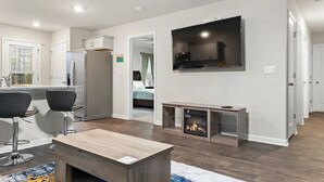 A modern living room with a flat-screen TV, a fireplace, and a kitchen area with a refrigerator and bar stools.