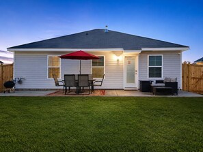 Une maison de plain-pied dotée d’un patio couvert comprenant une table et des chaises, un parasol rouge et un barbecue.