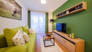 Uma sala de estar com um sofá verde, uma televisão de tela plana, um armário de madeira e um quadro de uma vaca na parede.