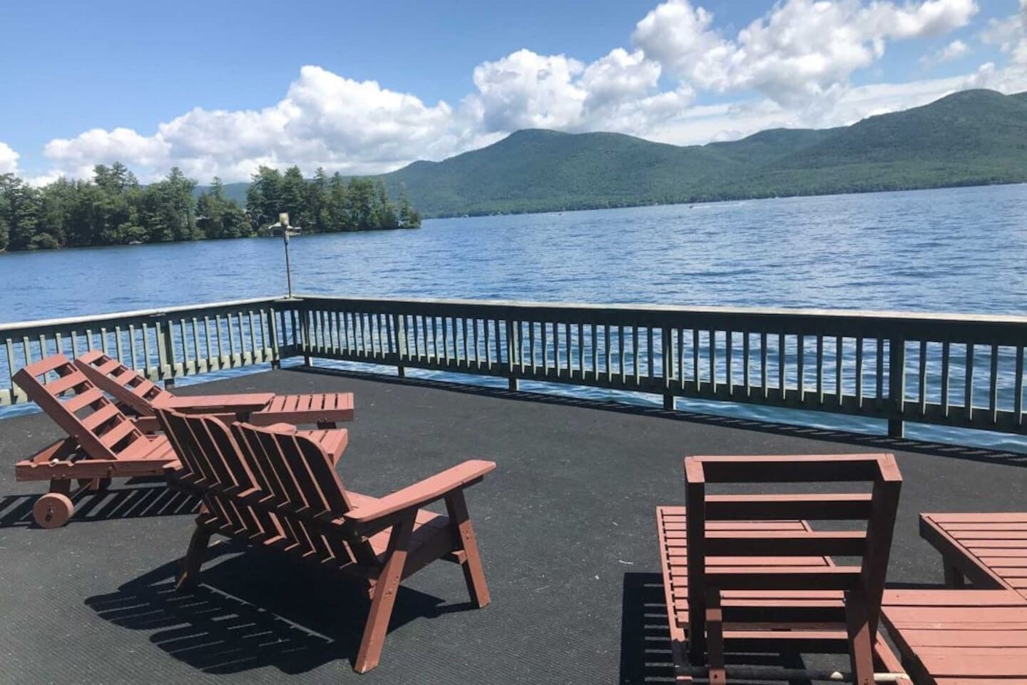 Una terraza con sillas y mesas de madera que da a un lago con montañas al fondo.