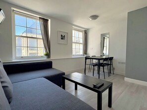 A modern living room with a sofa, a dining table with chairs, large windows with curtains, and a ceiling light. - Tottenham Flats by PrimeCityHosts 2F (Greater London)