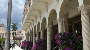 Un bâtiment orné d’une série d’arches et de colonnes, avec un palmier et des plantes en pot aux fleurs violettes.