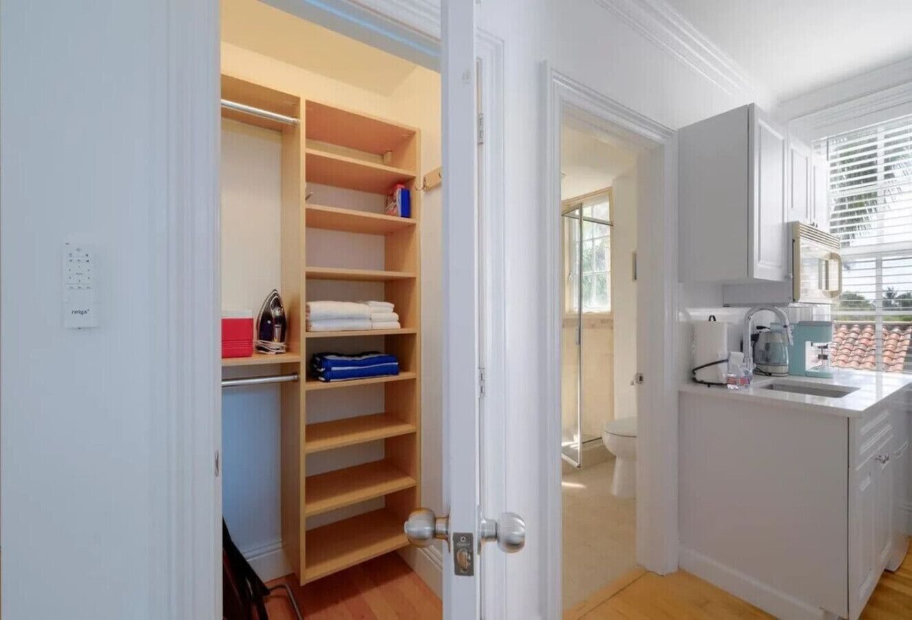 A modern kitchen with a built-in sink, a window with blinds, and a wooden door leading to a closet with shelves.