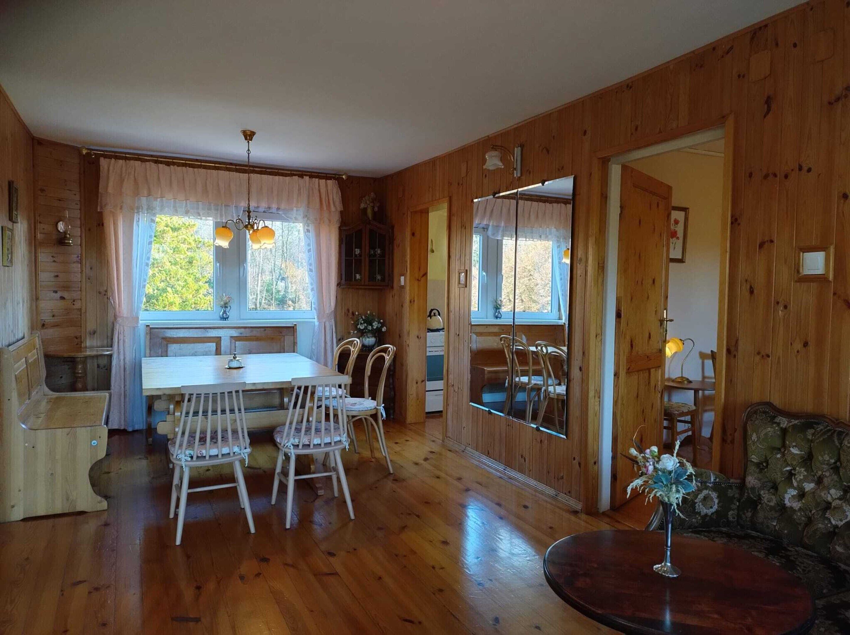 A cozy wooden cabin with a dining area featuring a table and chairs, a sofa, and a large window with curtains.