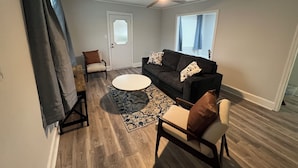 Un salon avec un canapé de couleur foncée, une table basse ronde et un tapis à motifs. On y trouve une porte avec une fenêtre, un ventilateur de plafond et un fauteuil avec un coussin.