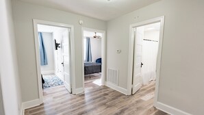Un couloir au sol en bois et aux murs blancs, qui mène à une chambre à coucher équipée d’un lit et d’un ventilateur de plafond.