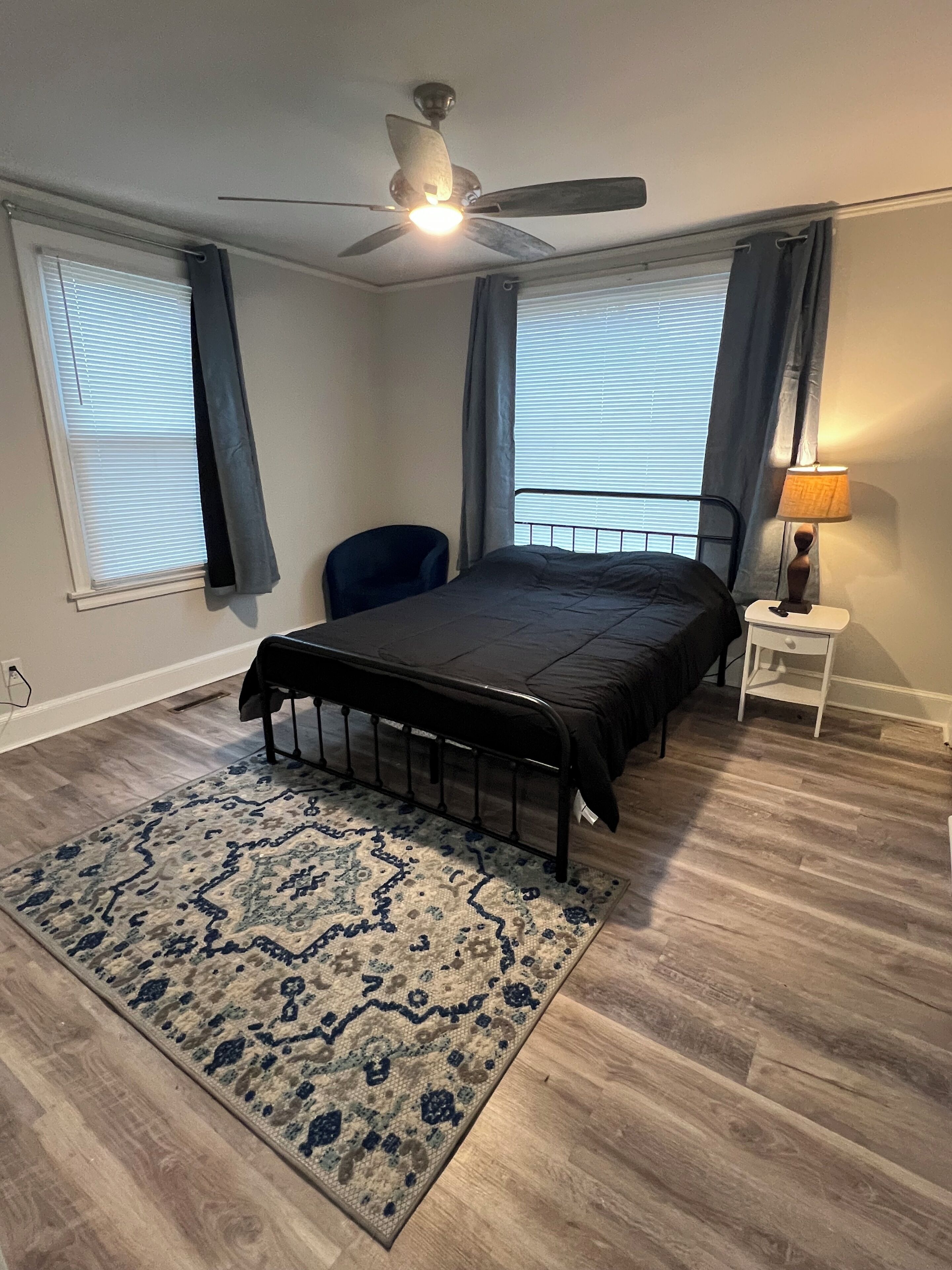 Quarto com cama, um ventilador de teto, mesa de cabeceira com abajur, cadeira e tapete estampado.