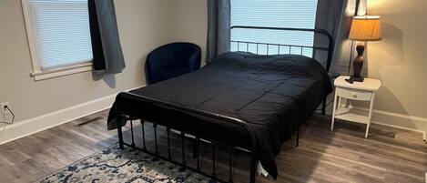 Quarto com cama, um ventilador de teto, mesa de cabeceira com abajur, cadeira e tapete estampado.