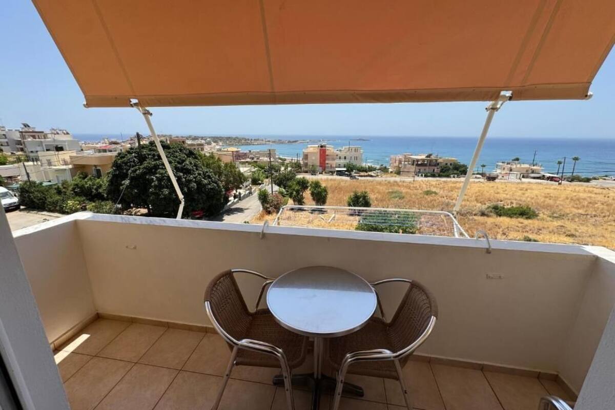 Ein Balkon mit Tisch und Stühlen, der Blick auf die Küste mit Gebäuden und dem Meer freigibt.