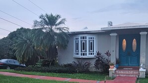 Ein Haus mit einem roten Weg, der zu einer blauen Tür führt, umgeben von Grünpflanzen und einer Palme.
