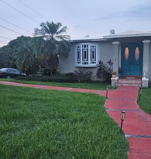 A house with a red pathway leading to a blue door, surrounded by greenery and a palm tree. - Diamond Stays Studio: Your quiet 1-Bdrm Caribbean Oasis, 8 min to Beach, Aguada (AGUADA)