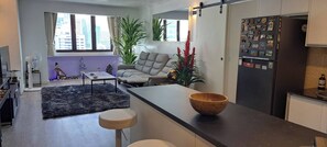 A modern kitchen with a black refrigerator adorned with magnets, a grey sofa, and a large window offering a city view. - Stylish New Condo in Nana (Bangkok)