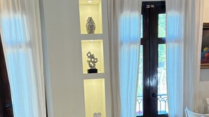 A room with a door, white curtains, a white chair, and a wall-mounted shelf with decorative items. - Casa de mabel (Santo Domingo)