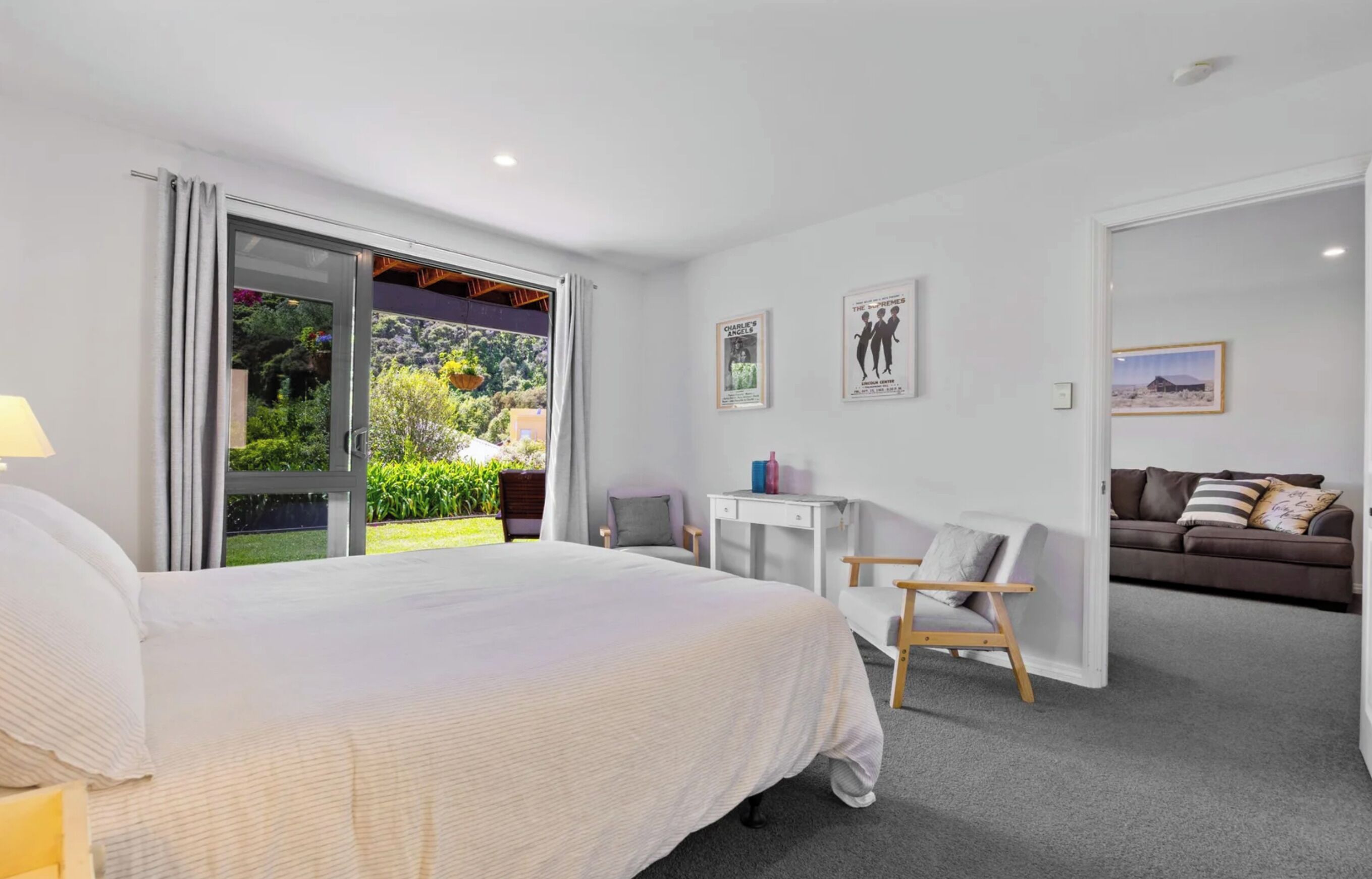 A bedroom with a large bed, a desk, and a sofa. There is a sliding glass door leading to a balcony with a view of greenery.